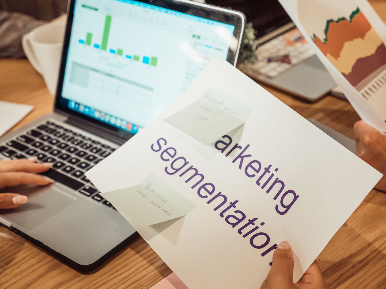 A contemporary workspace where hands review marketing data on a laptop alongside paper documents.